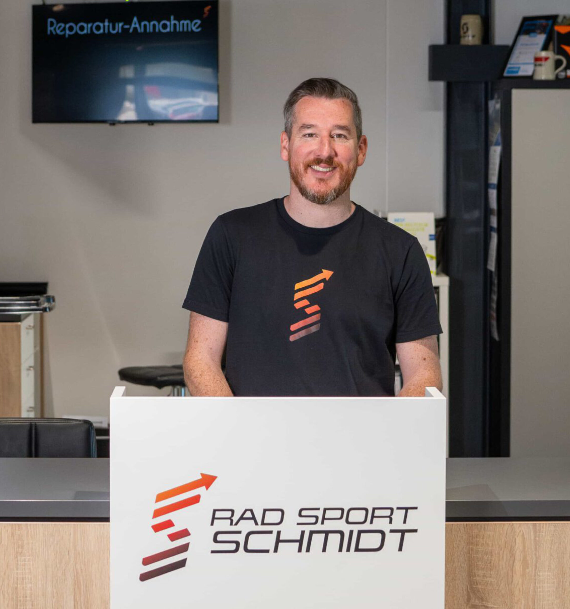 Ein lächelnder Mann mit kurzen Haaren und Bart steht hinter einem weißen Tresen mit dem Logo von RAD SPORT SCHMIDT. Über ihm steht ein Schild mit der Aufschrift „Reparatur-Annahme“. Es scheint sich um einen Fahrradladen oder eine Reparaturwerkstatt zu handeln.