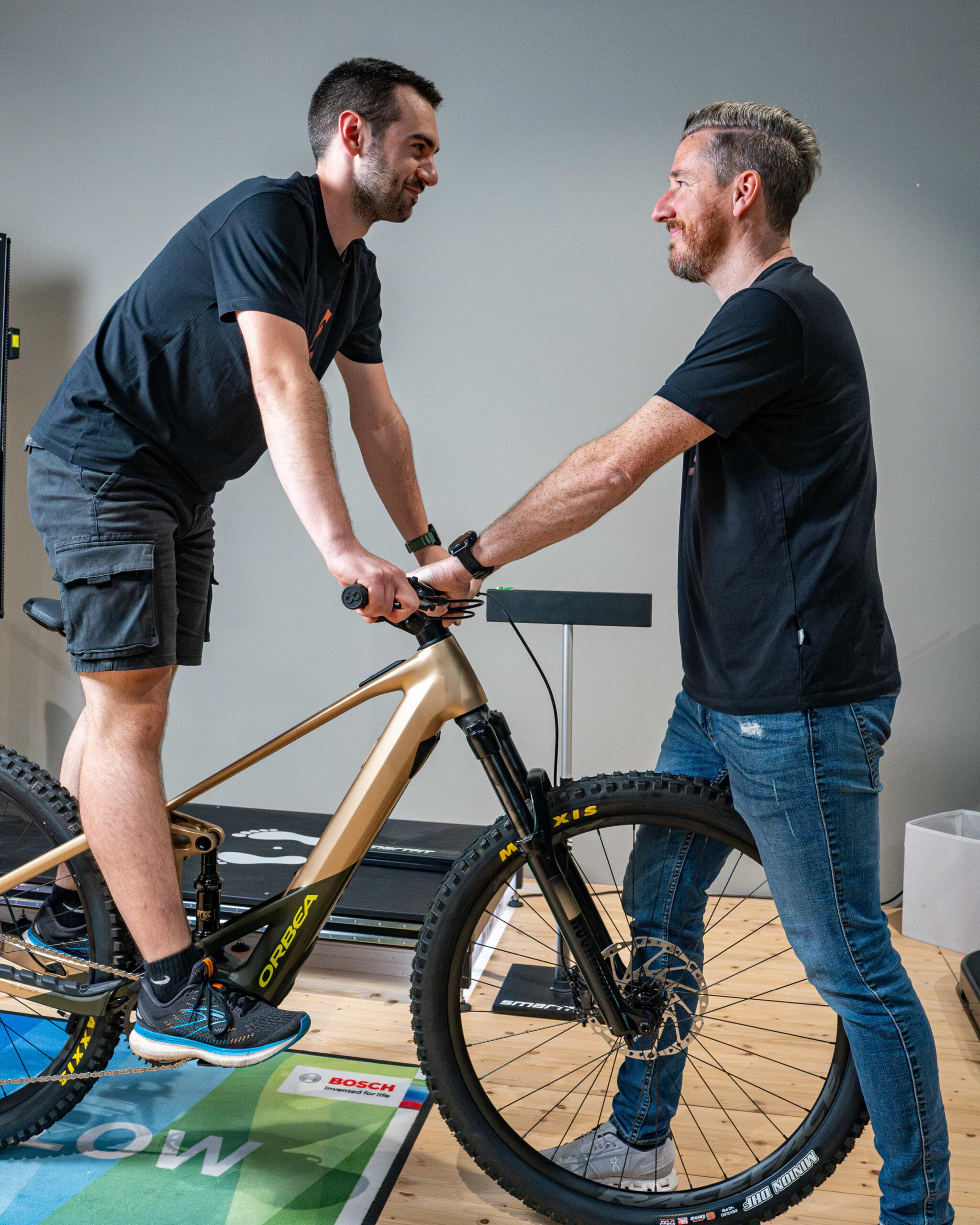 Zwei Männer lächeln sich an, während einer auf einem goldenen Orbea-Mountainbike sitzt und der andere drinnen auf einem Holzboden steht und den Lenker festhält.