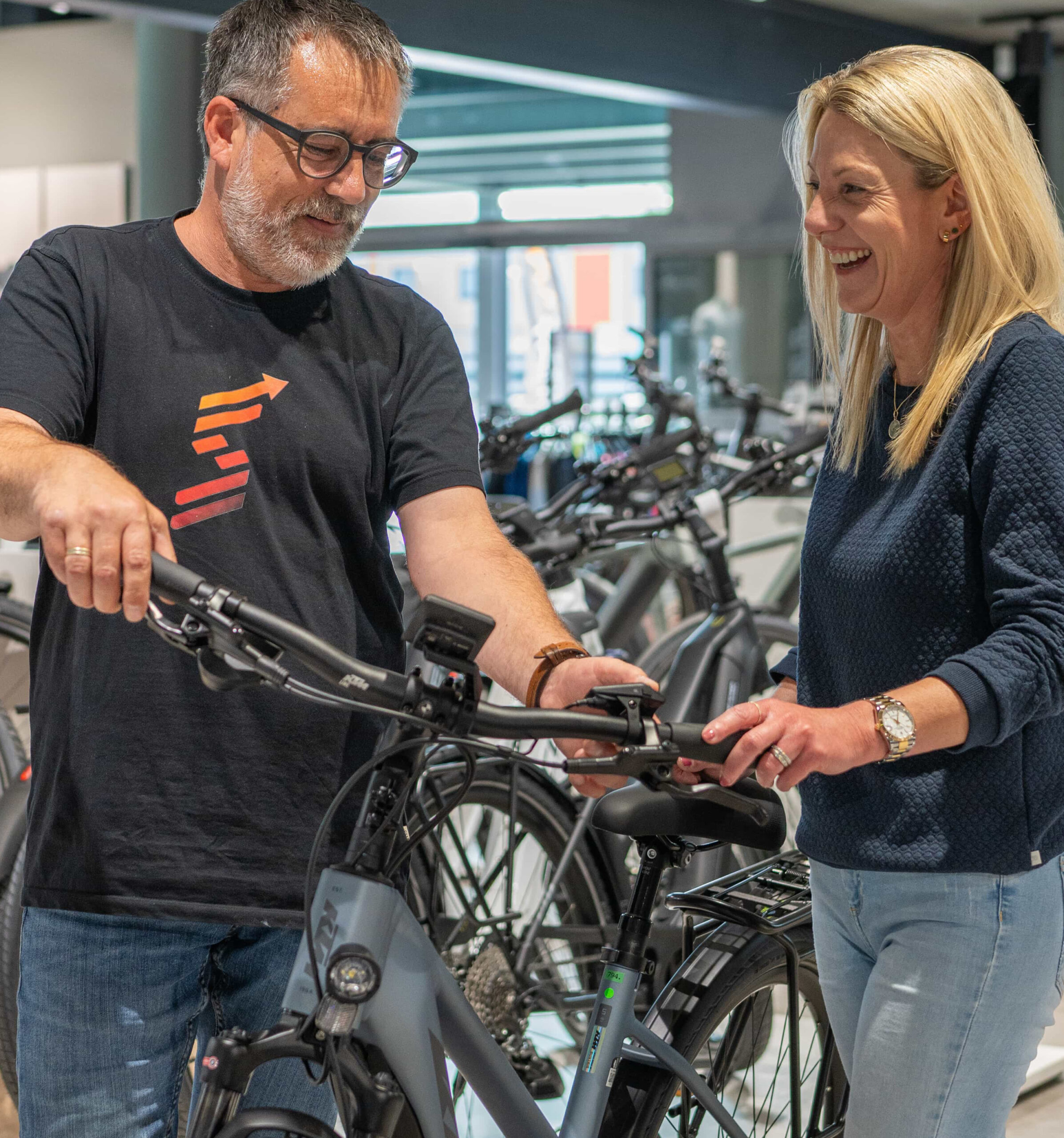 Ein Mann und eine Frau stehen lächelnd neben einem Fahrrad in einem Fahrradladen. Der Mann zeigt auf den Lenker des Fahrrads, im Hintergrund sind weitere Fahrräder zu sehen.