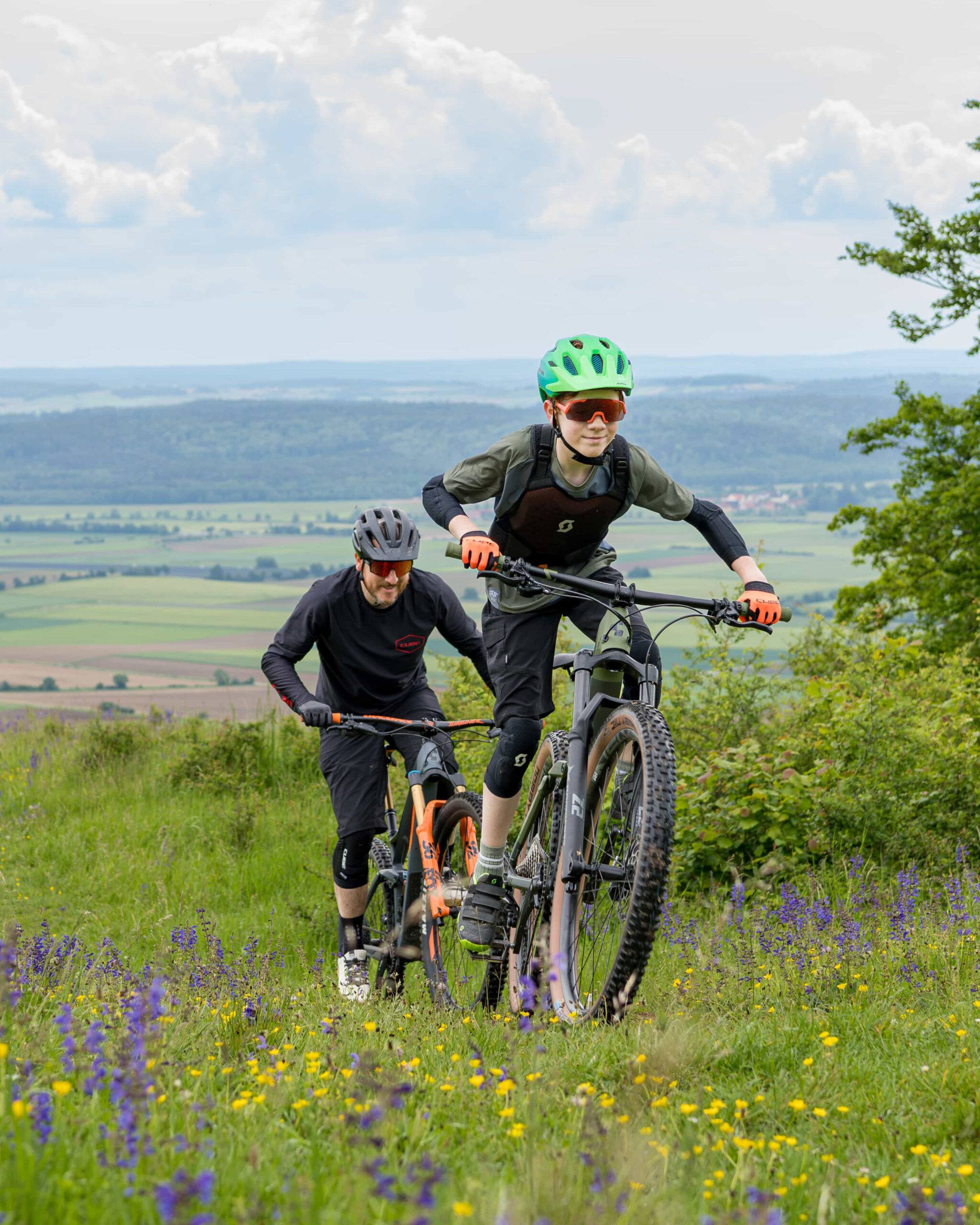 Zwei Personen fahren mit Mountainbikes bergauf durch eine grasbewachsene, blumenreiche Landschaft. Im Hintergrund sind Hügel und Felder zu sehen. Der Vordermann trägt einen grünen Helm und eine Sonnenbrille. Beide sind sportlich gekleidet.