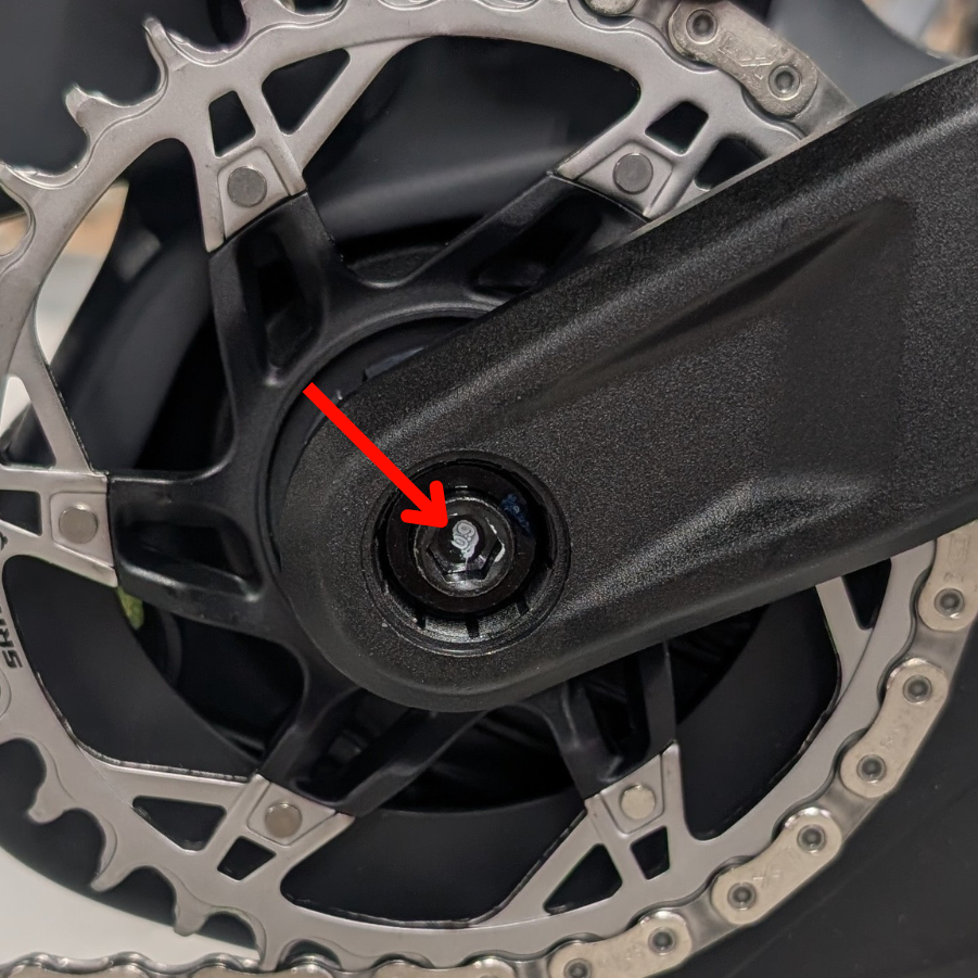 AcidKurbel_Rckruf_Abb2_MarkierungAntrieb_web Nahaufnahme eines Fahrradkurbelarms und eines Kettenblatts mit einem roten Pfeil, der auf eine Schraube in der Mitte des Kurbelarms zeigt.