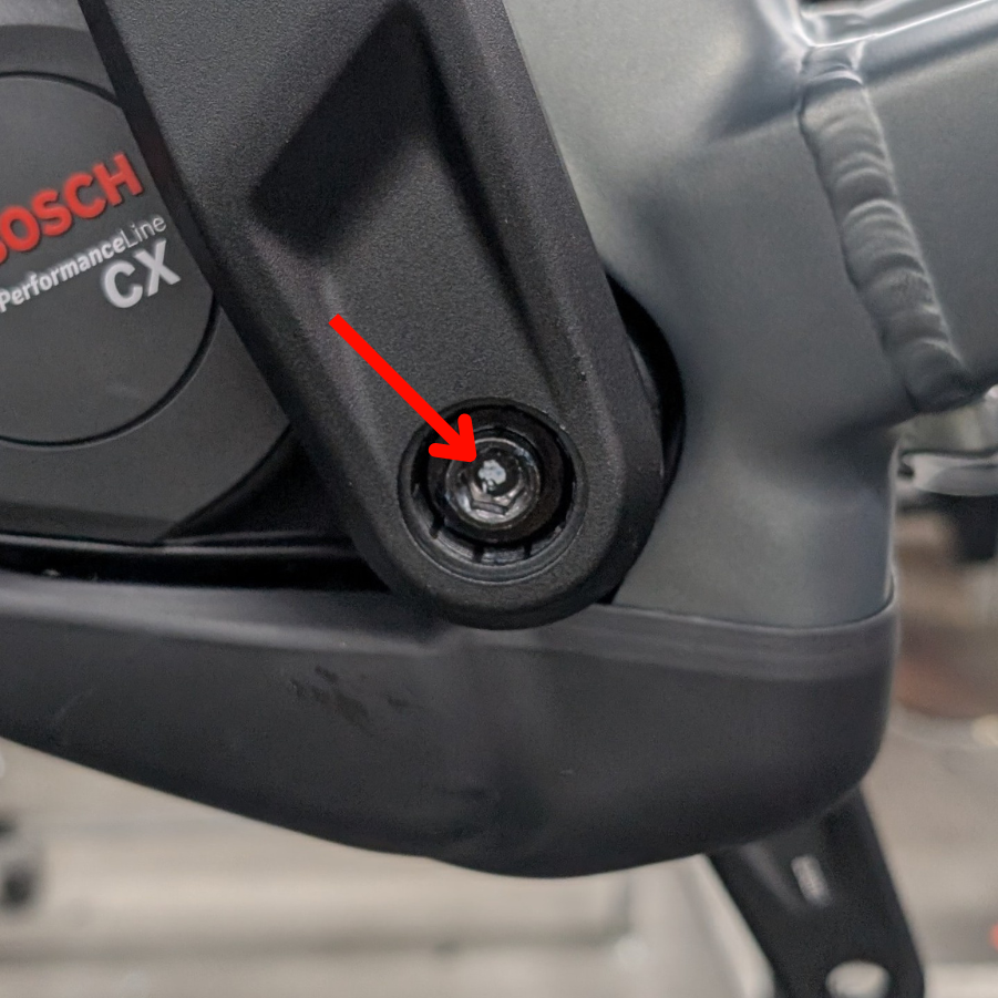 AcidKurbel_Rckruf_Abb3_MarkierungAntrieb_web Nahaufnahme eines Bosch Performance Line CX-Elektroradmotors mit einem roten Pfeil, der auf eine glänzende, silberne Schraube in einer schwarzen, runden Aussparung am Motorgehäuse zeigt.